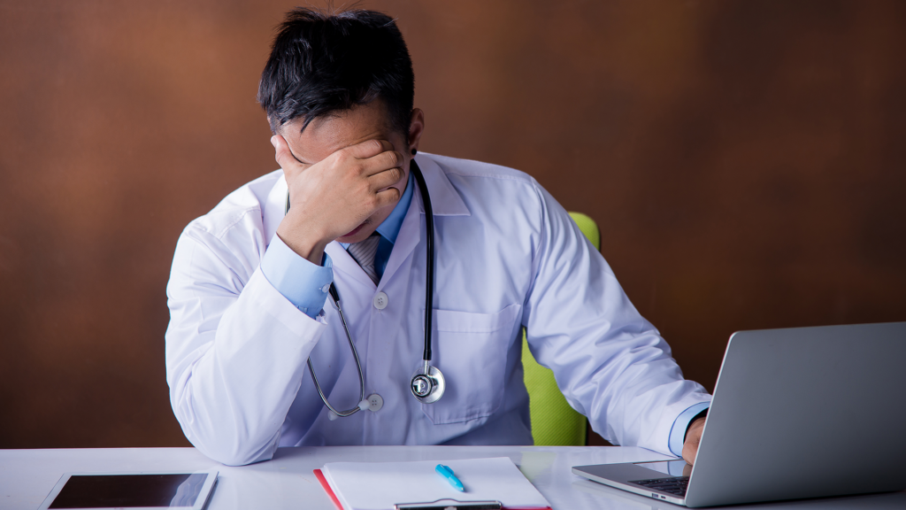 Doctor sitting at a desk with hand on face, showing signs of fatigue and stress in a professional setting